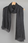 A black, lightweight, crinkled Color-POP Scarf from looselyboho is draped over a wooden hanger. Perfect for all-weather wear, the scarf has a fringed edge and appears to be made of a soft, airy fabric. The background is plain gray.