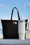 A black tote bag with a white monogram "W" sits next to a tall, spill-proof Indestructible Travel Cup by looselyboho with a handle and straw on a concrete surface, against a blurry beach background.