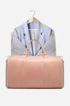 A light pink Smart No-Wrinkle Foldable Travel Bag by The Windbreaker Jacket is positioned in the foreground, featuring waterproof PU leather. In the background, a light blue blazer hangs on a wooden hanger. The image has a minimalistic setup with a plain white backdrop.