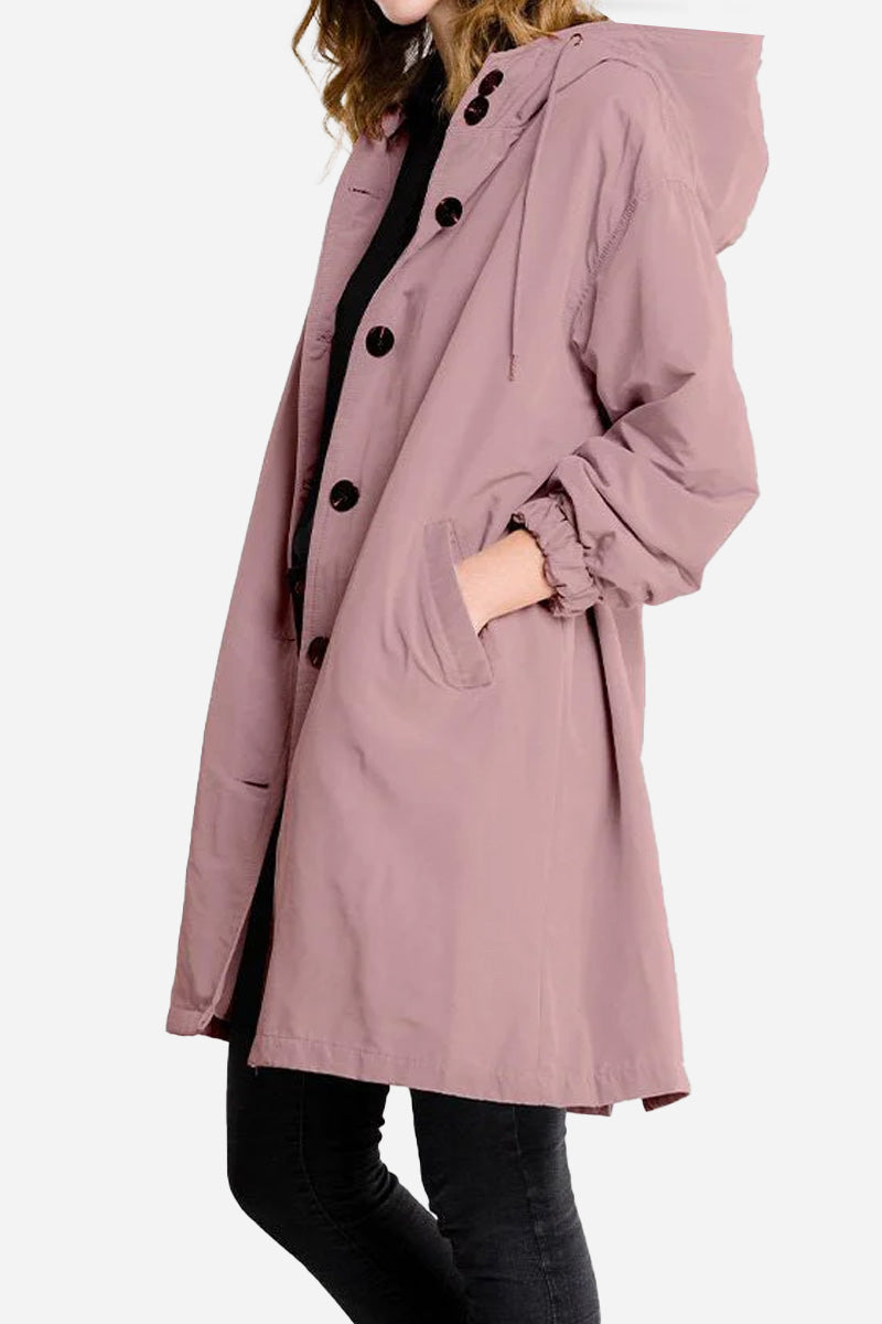 A person wearing The WJacket's Water Resistant Oversized Hooded Windbreaker Rain Jacket in light purple with black buttons and front pockets. This timelessly stylish jacket with its rare beautiful color features long sleeves, and the person is standing sideways with their left hand in the pocket. The background is plain white.