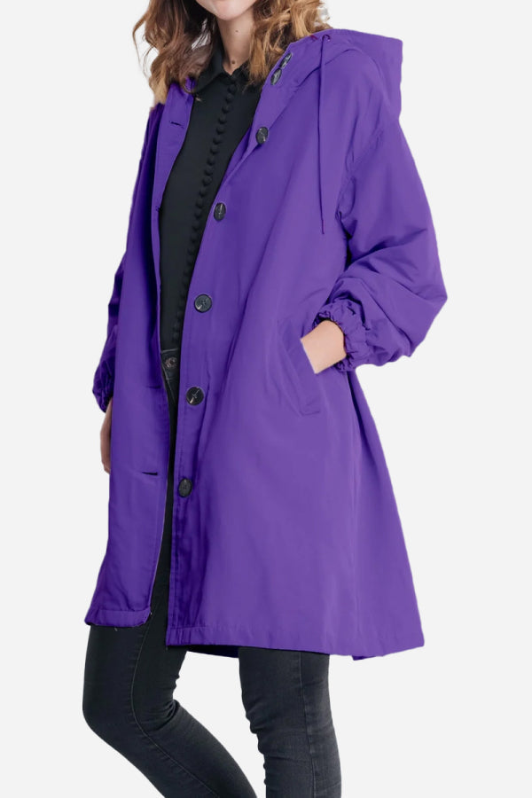 A person is wearing a Water Resistant Oversized Hooded Windbreaker Rain Jacket by The WJacket in purple with black buttons over a black outfit. Embodying timeless style, the jacket features pockets where the person's hands are tucked away. The background is plain white.