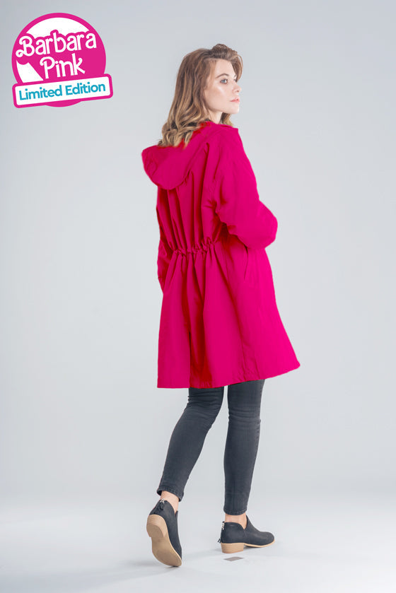 A woman stands looking over her shoulder, wearing a bright pink Water Resistant Oversized Hooded Windbreaker Rain Jacket from The WJacket. She is also dressed in dark pants and black shoes. Text on the image reads: 