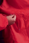 A person is adjusting the fit of a red Water Resistant Oversized Hooded Windbreaker Rain Jacket by pulling its matching drawstring. The person's hand is partially visible, highlighting the timeless style and versatility of The WJacket.