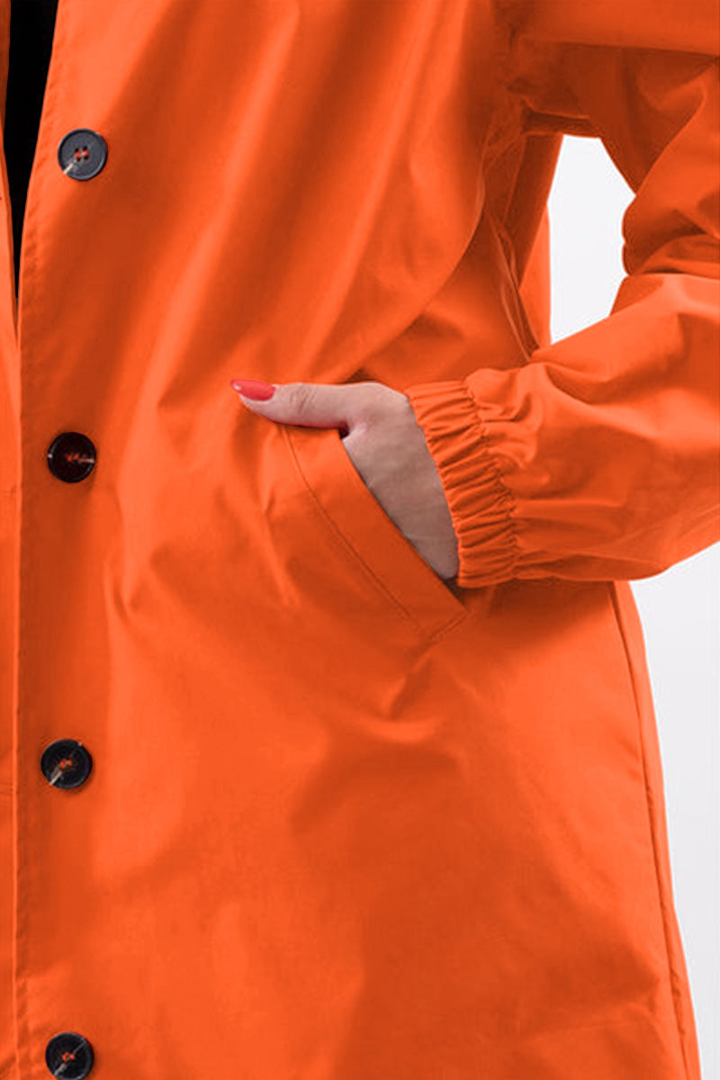 A person is shown wearing the looselyboho PUMPKIN ORANGE WINDBREAKER, featuring black buttons. Their hand is partially tucked into one of the jacket's pockets. This lightweight coat also has elastic at the wrists, and the background is plain white.