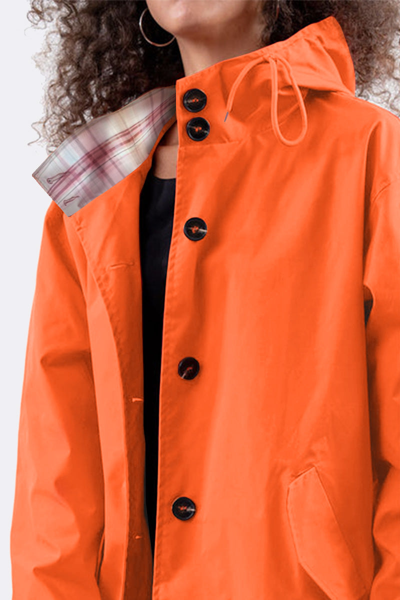 A person dressed in a looselyboho PUMPKIN ORANGE WINDBREAKER, which has large black buttons and a plaid lining. This lightweight jacket includes a drawstring hood. The individual's face is not visible, and they have curly hair and hoop earrings.