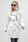 A woman with long, blonde hair is wearing a BELTED FROST WHITE WINDBREAKER by looselyboho, black sunglasses, and dark jeans. She stands against a plain gray background, with one hand holding the front of her coat. She wears a white top underneath and layered necklaces.