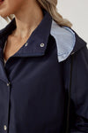 Close-up of a person wearing the STRIPED NAVY SHADOW WINDBREAKER JACKET by looselyboho with silver button fastenings and a zipper. The collar is folded down, revealing a blue and white striped lining on the inside. The person's head is partially visible, showing blonde hair.