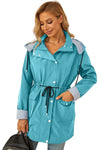 A woman is wearing a teal, button-up waterproof STRIPED TURQUOISE OASIS WINDBREAKER JACKET from looselyboho with adjustable drawstrings at the waist and hood. The coat features front pockets and striped inner lining on the cuffs and hood. She has blonde hair and is looking down, holding a black bag in one hand.