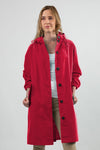 A woman with long blonde hair is wearing a bright red, knee-length VIVA MAGENTA WINDBREAKER by looselyboho with black buttons and a hood. She has a white top underneath and beige pants. The background is plain and light gray. She is looking slightly to the side with a neutral expression.