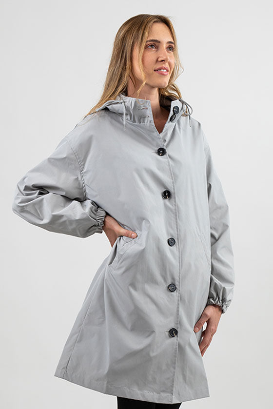 A woman with long, wavy hair is wearing a looselyboho ICE GRAY WINDBREAKER with black buttons made from water-resistant material. She is standing against a plain white background, with her left hand in her pocket and her right hand resting on her hip, looking slightly towards the right.