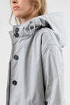 A person wearing a looselyboho ICE GRAY WINDBREAKER made from water-resistant material with black buttons. The focus is on the upper part of the windbreaker, highlighting the details of the adjustable hood and buttons. The background is plain and minimalistic.