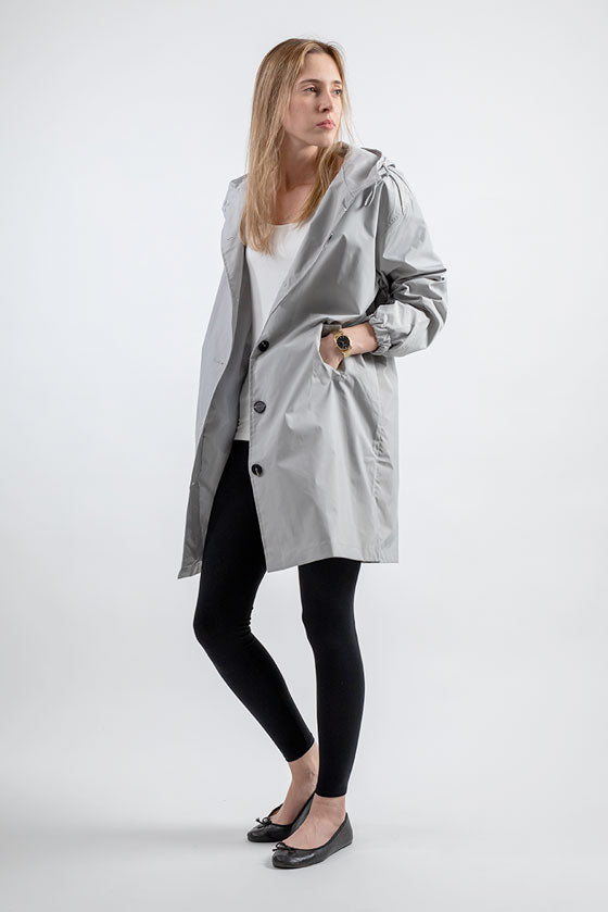 A woman with long blonde hair stands against a plain white background. She is wearing the ICE GRAY WINDBREAKER from looselyboho made from water-resistant material over a white top, black leggings, and black ballet flats. Her body is turned slightly to the side, and her hands are in the pockets of her coat.