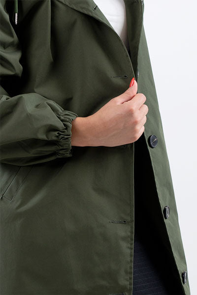 A person with a visible hand featuring red nail polish is buttoning up an OLIVE GREEN WINDBREAKER from The Windbreaker Jacket, which is lightweight, water-resistant, and has an elastic cuff on the sleeve. The jacket also has an adjustable hood. The person is wearing a white top underneath.