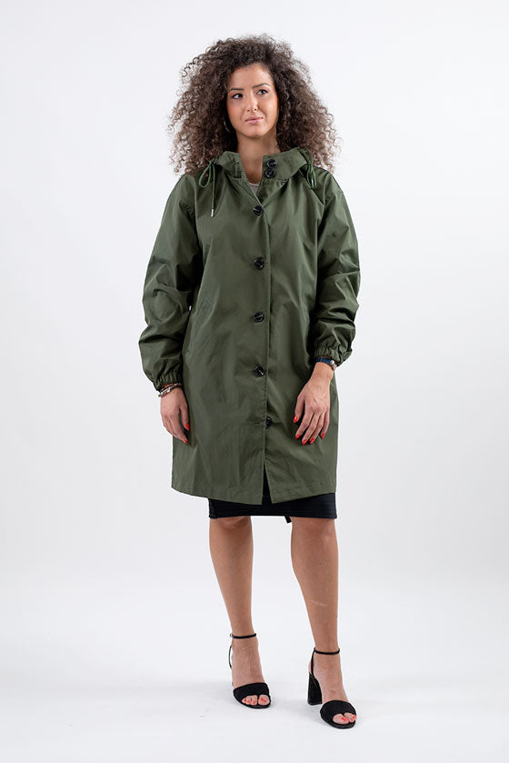 A woman with curly hair is wearing an OLIVE GREEN WINDBREAKER from The Windbreaker Jacket and a black skirt. She is standing on a white background, slightly smiling with one hand by her side and the other in her pocket. She is also wearing black high-heeled sandals.