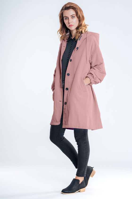 A person with long hair is wearing The Windbreaker Jacket's PASTEL PINK WINDBREAKER, a black top, slim dark jeans, and black ankle boots with a wooden heel. They are standing against a plain white background, showcasing their stylish clothing.